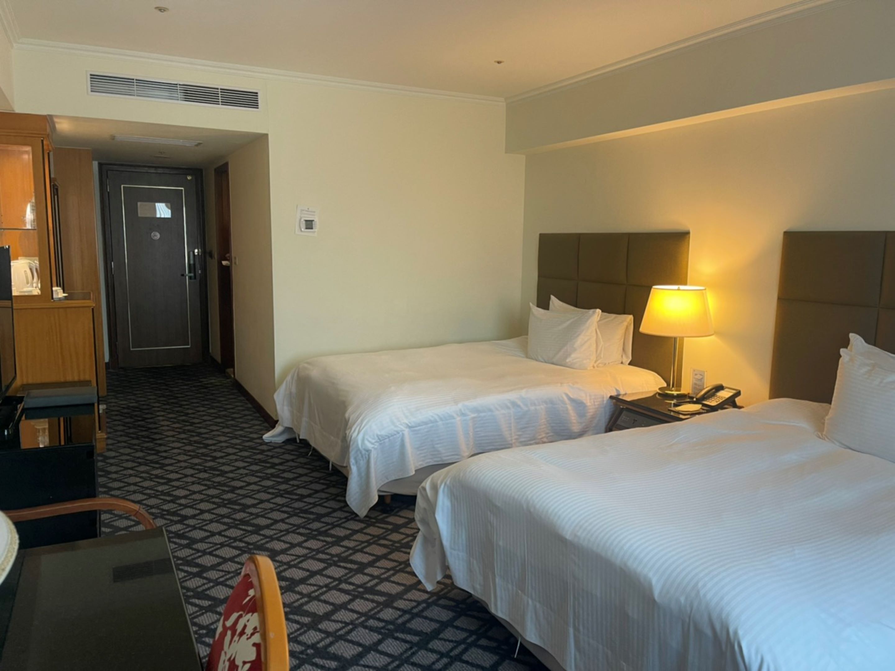 A hotel room with two beds, a desk, a chair, and a lamp.