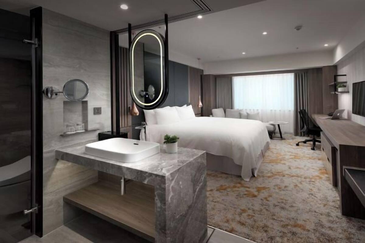 A modern hotel room with a marble bathroom, a bed with white linens, a desk, and a large window.