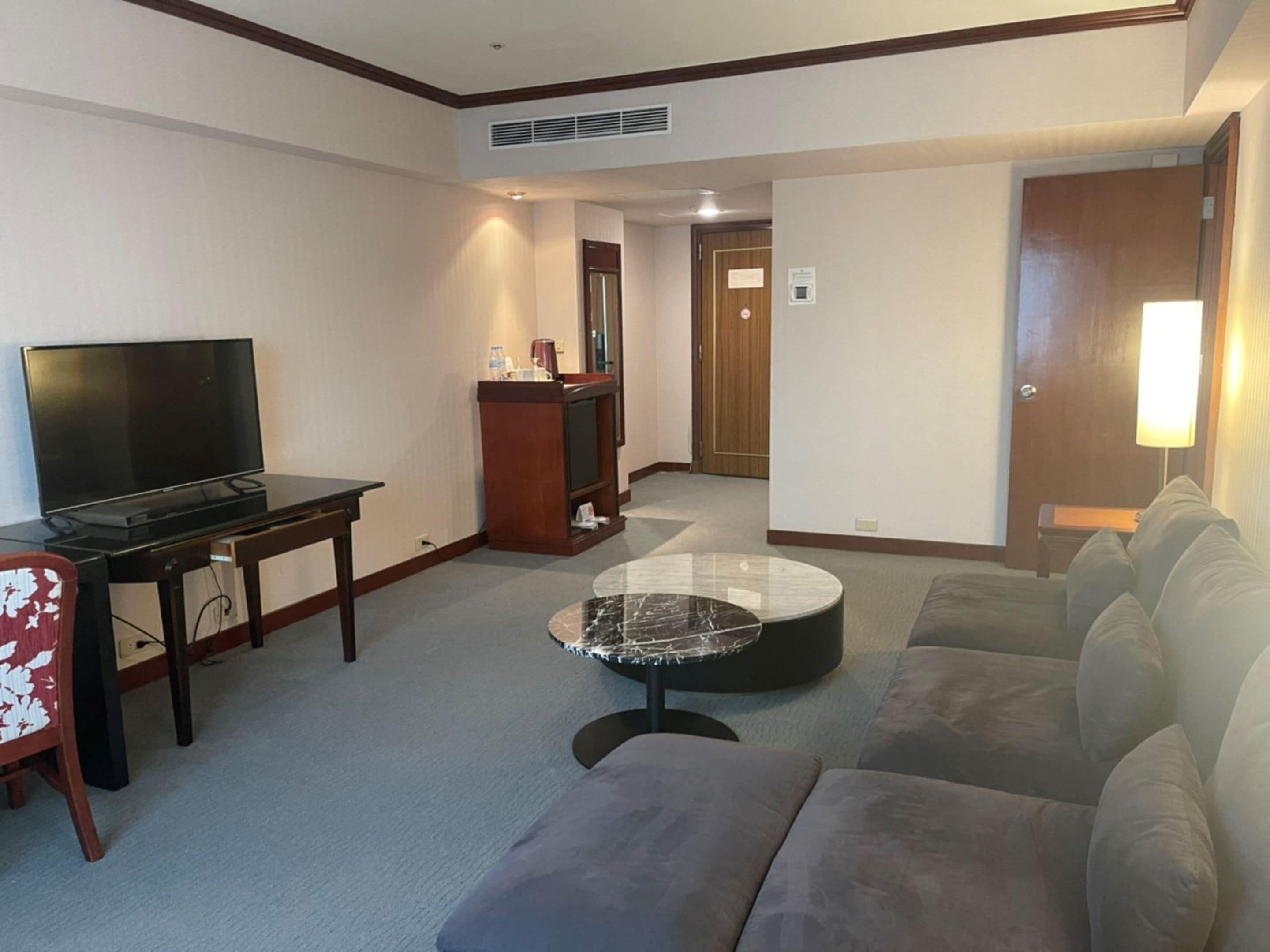 A hotel room with a flat-screen TV, a wooden desk, a marble coffee table, a grey sofa, and a wooden door.