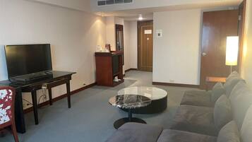 A hotel room with a flat-screen TV, a wooden desk, a marble coffee table, a grey sofa, and a wooden door.