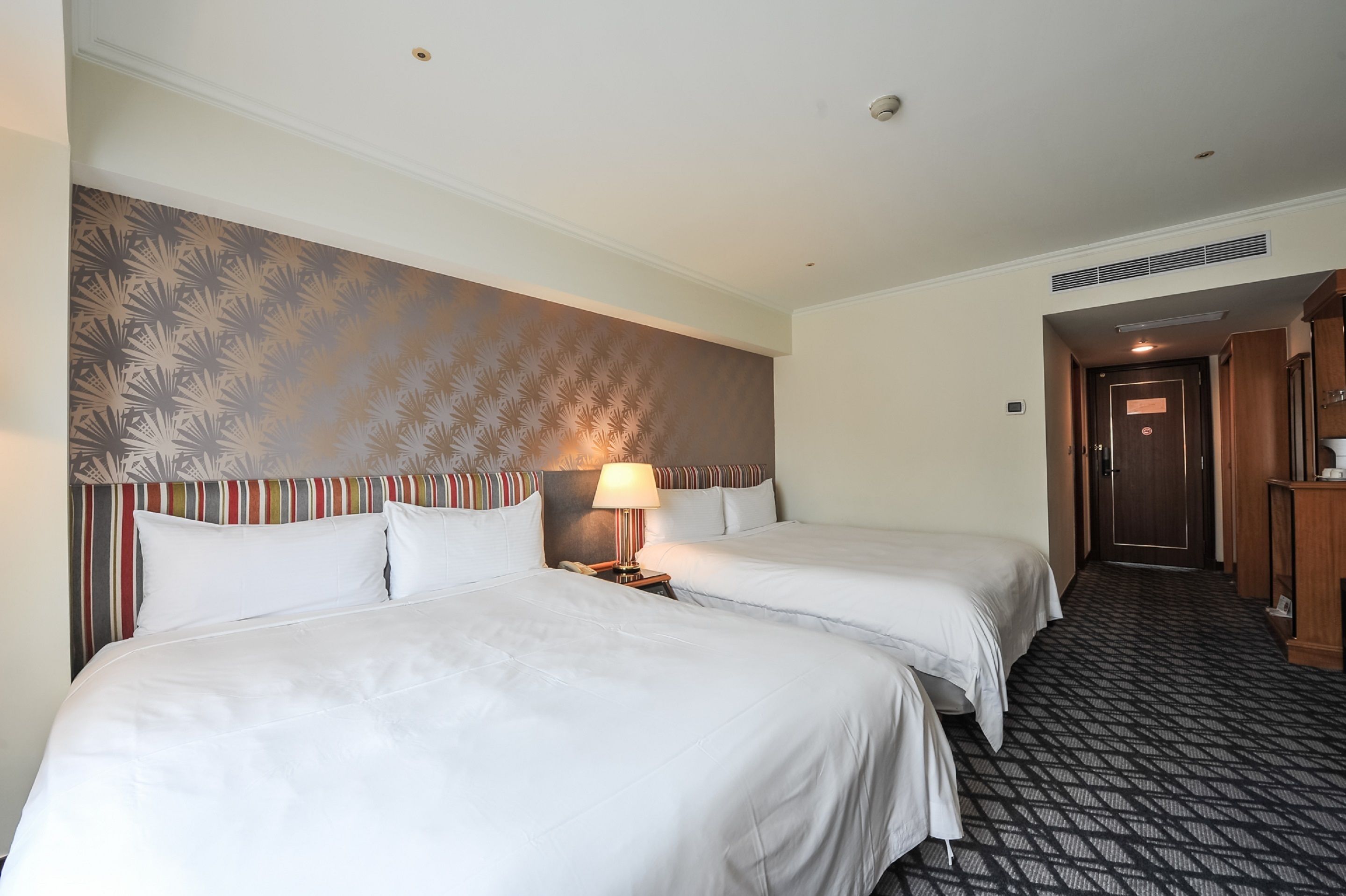 A hotel room with two beds, a striped headboard, a patterned wall, a lamp, and a carpeted floor.