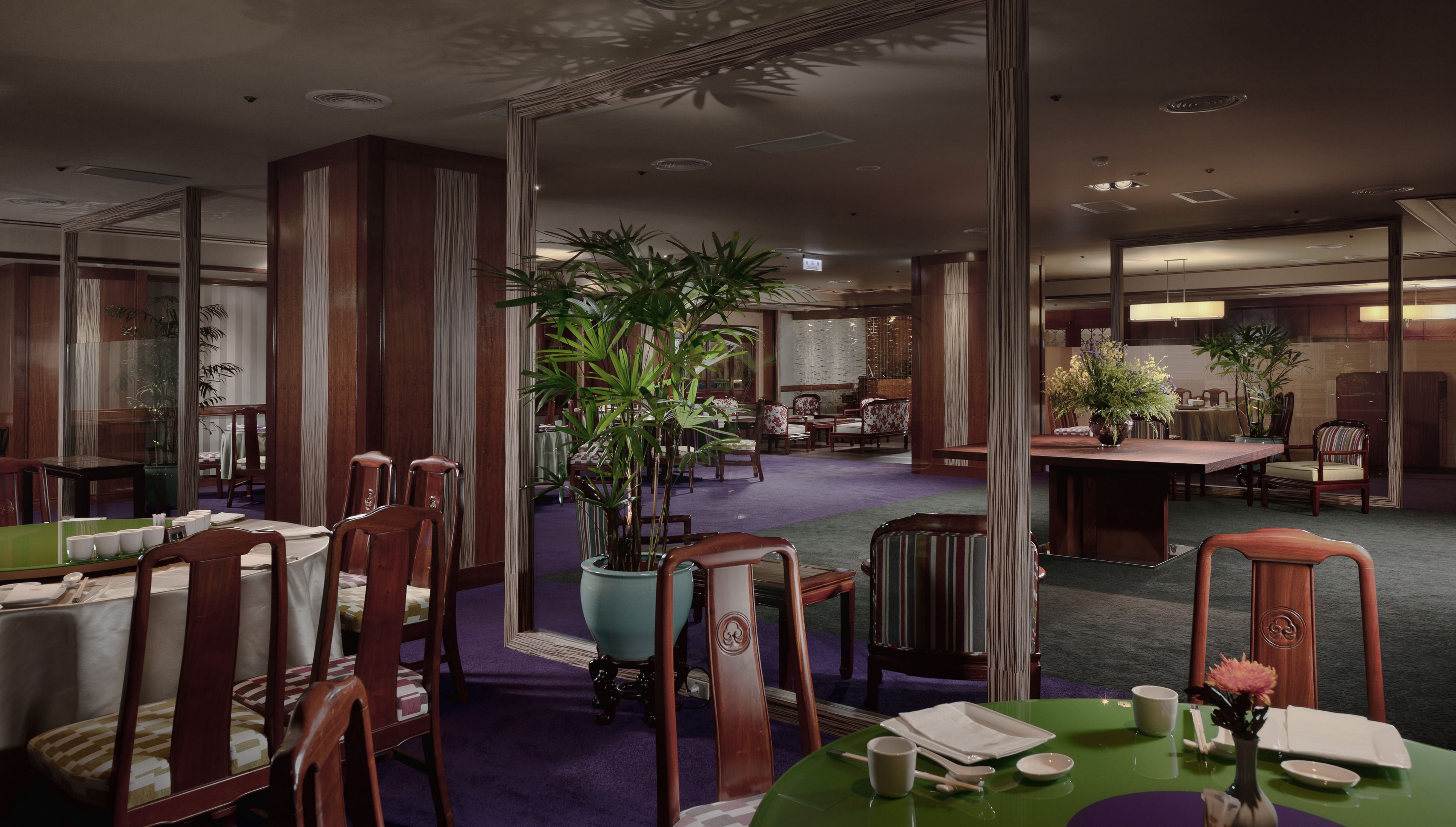 A spacious hotel dining area with multiple tables and chairs, large potted plants, and a high ceiling with recessed lighting.