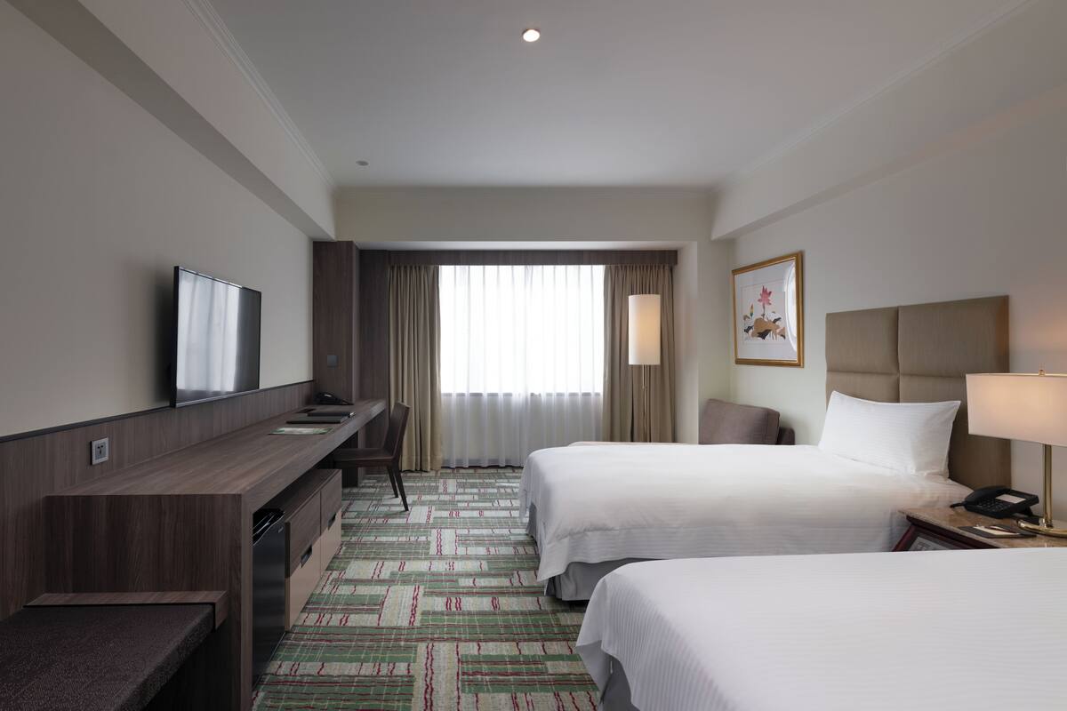 A hotel room with two beds, a desk, a TV, a lamp, and a window with curtains.