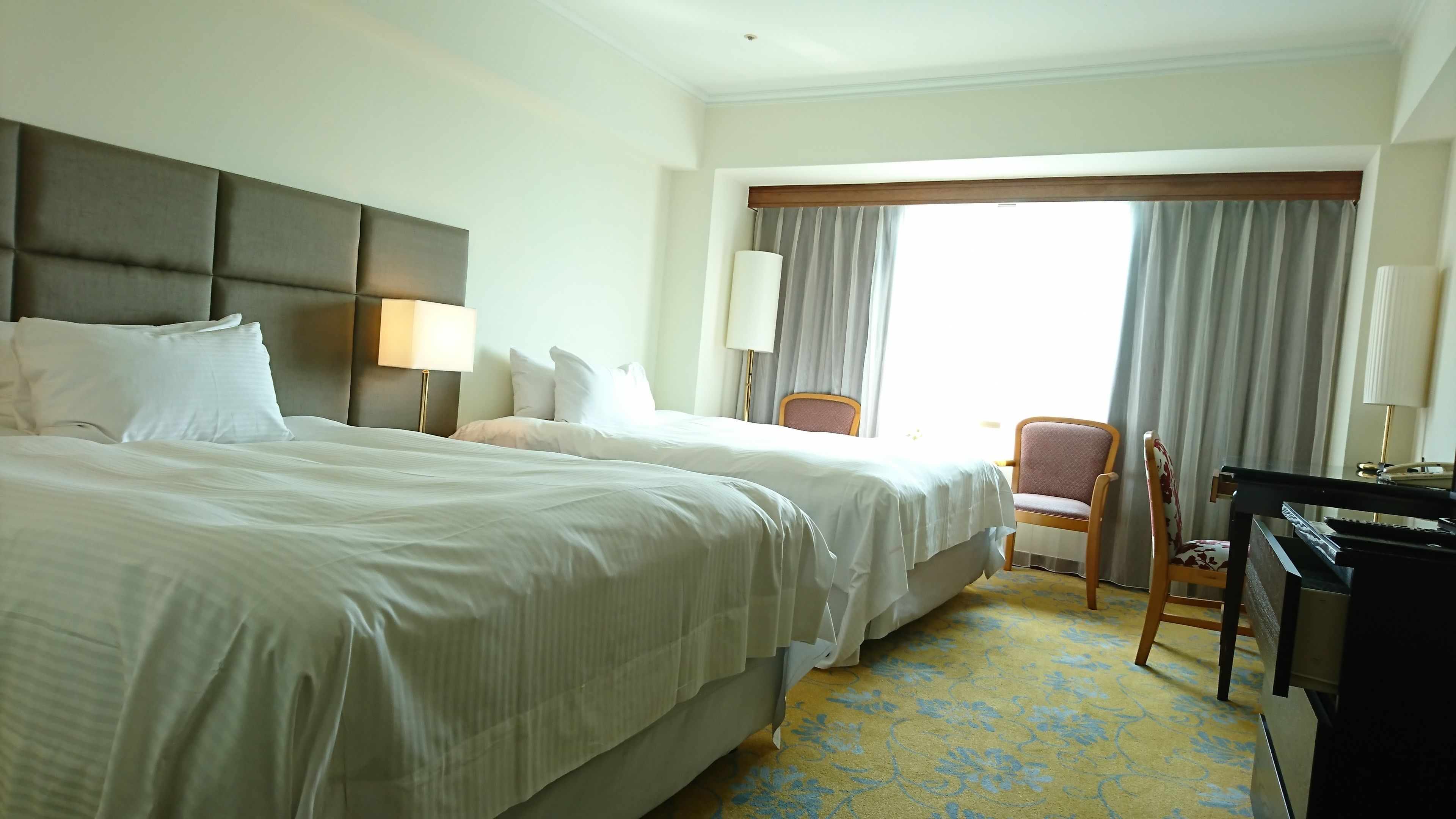 A hotel room with two beds, a desk, a chair, and a large window with curtains.