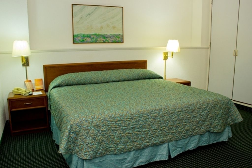 Una habitación de hotel con una cama, dos lámparas de noche, una mesita de noche con un teléfono y un cuadro en la pared.