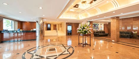 A spacious lobby with a polished floor, a reception desk, a staircase, and a seating area with a floral arrangement.