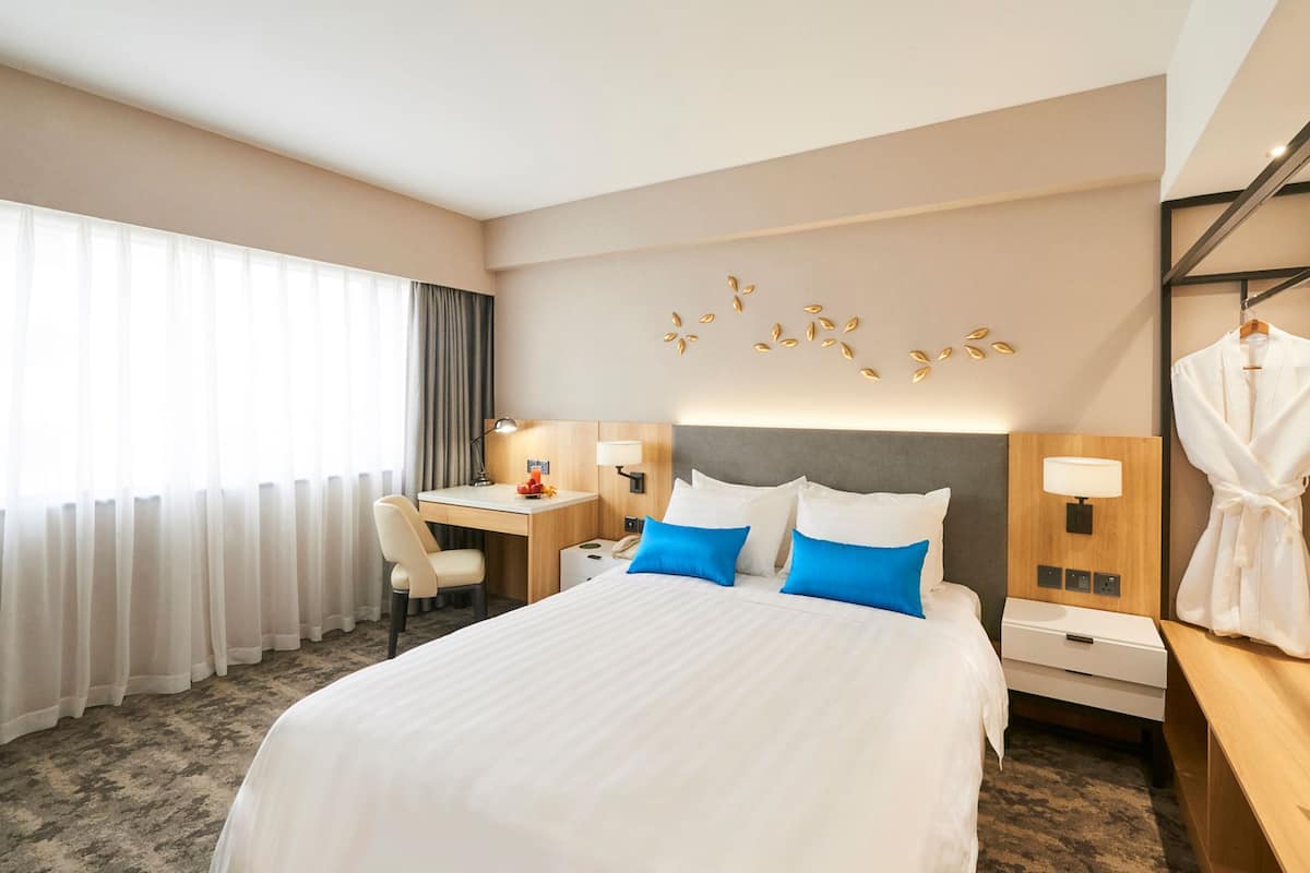 A hotel room with a large bed, a desk, a chair, a robe rack, and a wall-mounted lamp.