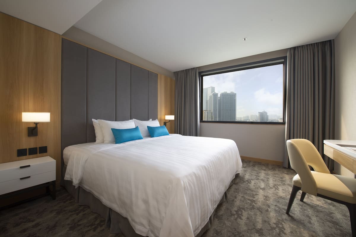 A hotel room with a large bed, a desk, and a view of the city skyline through a large window.