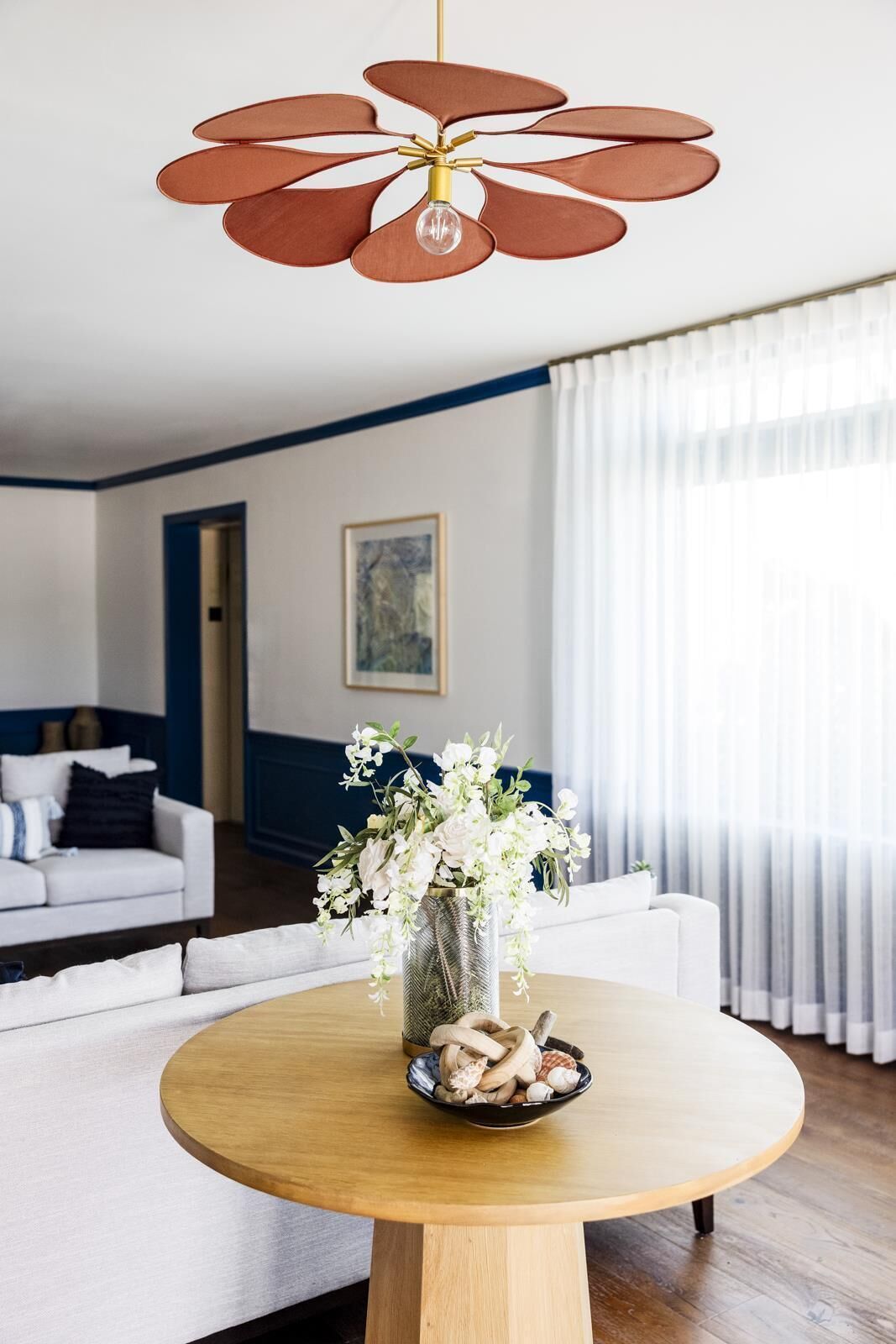 Ein modernes Wohnzimmer mit einem runden Holztisch, einer Vase mit weißen Blumen und einem Deckenventilator mit braunen Flügeln.