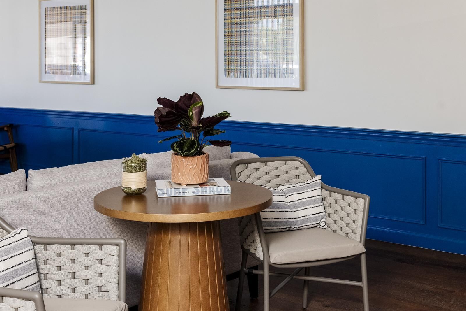 Een ronde houten tafel met een bloempot en een boek met de titel 'SURF SHACK' in een kamer met blauwe lambrisering en twee ingelijste kunstwerken aan de muur.