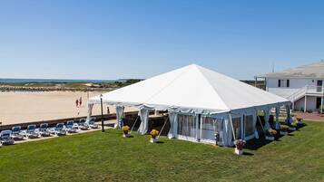 Uma tenda à beira-mar com um toldo branco, cercada por cadeiras de grama e plantas em vasos, com vista para a praia.