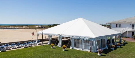 Una carpa junto a la playa con dosel blanco, rodeada de sillas de jardín y plantas en macetas, con vista al mar.
