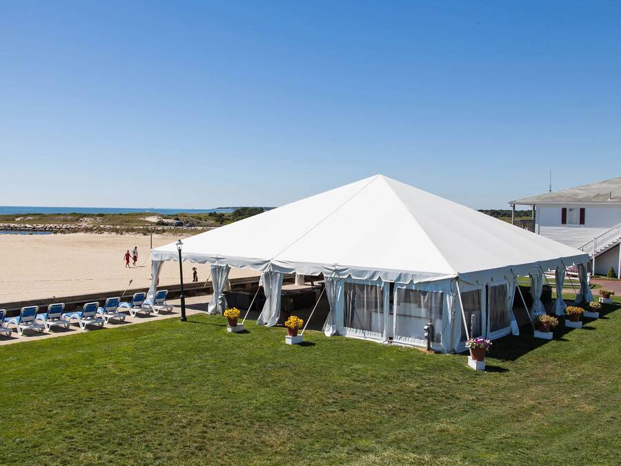 Uma tenda à beira-mar com um toldo branco, cercada por cadeiras de grama e plantas em vasos, com vista para a praia.