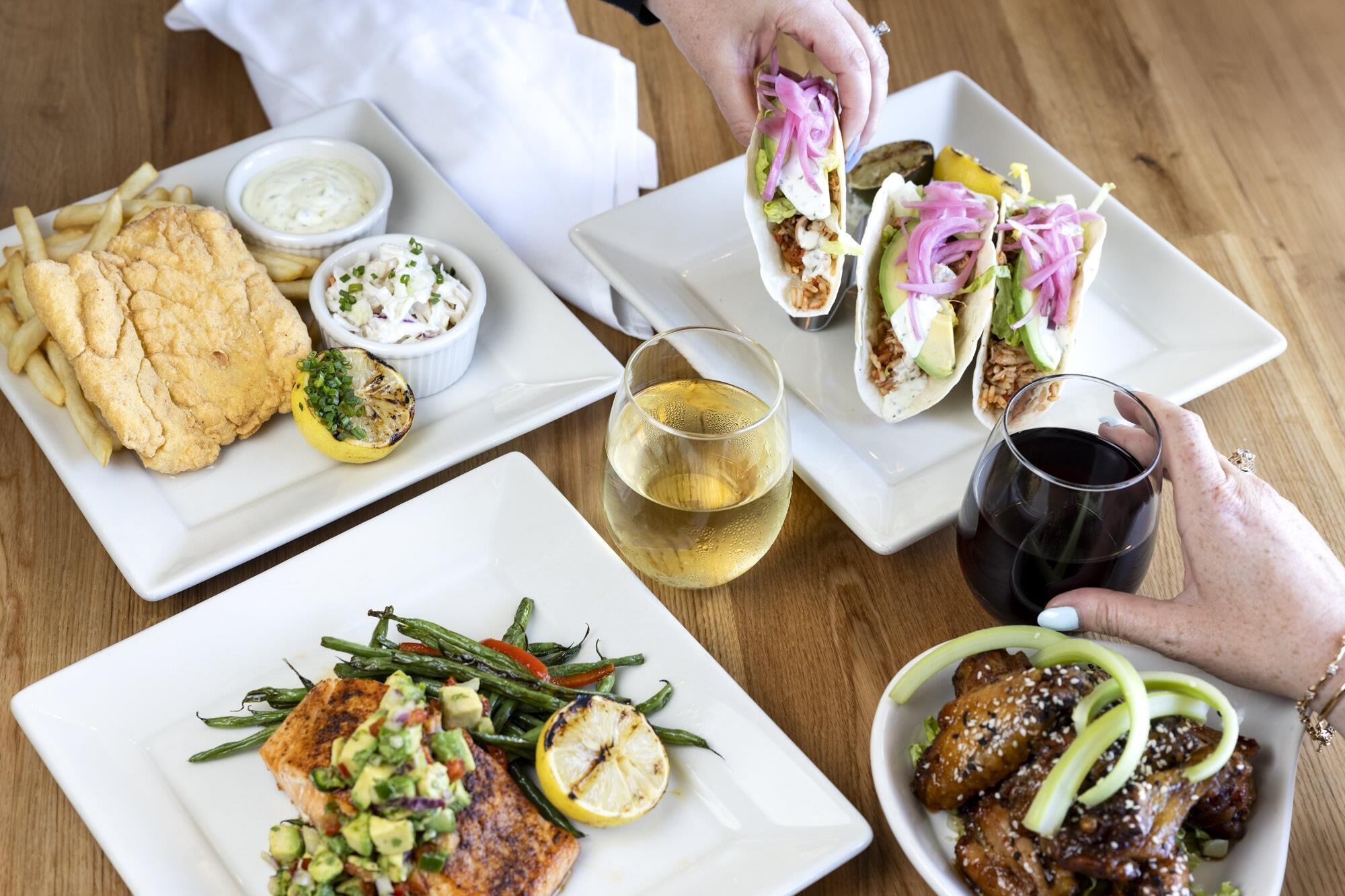 A variety of dishes including fish, tacos, and a salad, along with two glasses of wine on a wooden table.