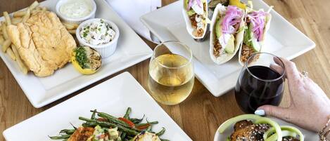 A variety of dishes including fish, tacos, and a salad, along with two glasses of wine on a wooden table.