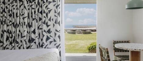 A bedroom with a bed, a view of the sea, a dining area with a table and chairs, and a patterned curtain.