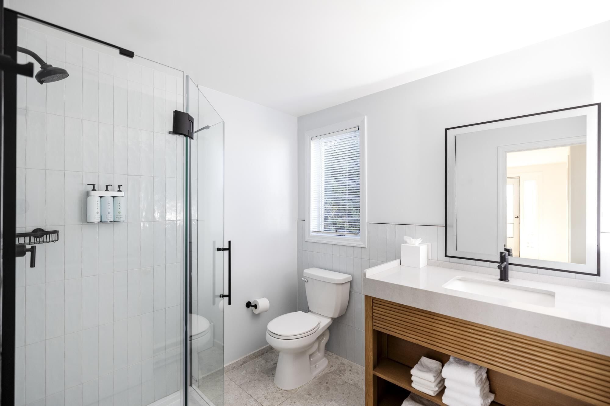 Une salle de bain moderne avec une douche à parois de verre, des toilettes, un lavabo avec un miroir et une fenêtre équipée de stores.