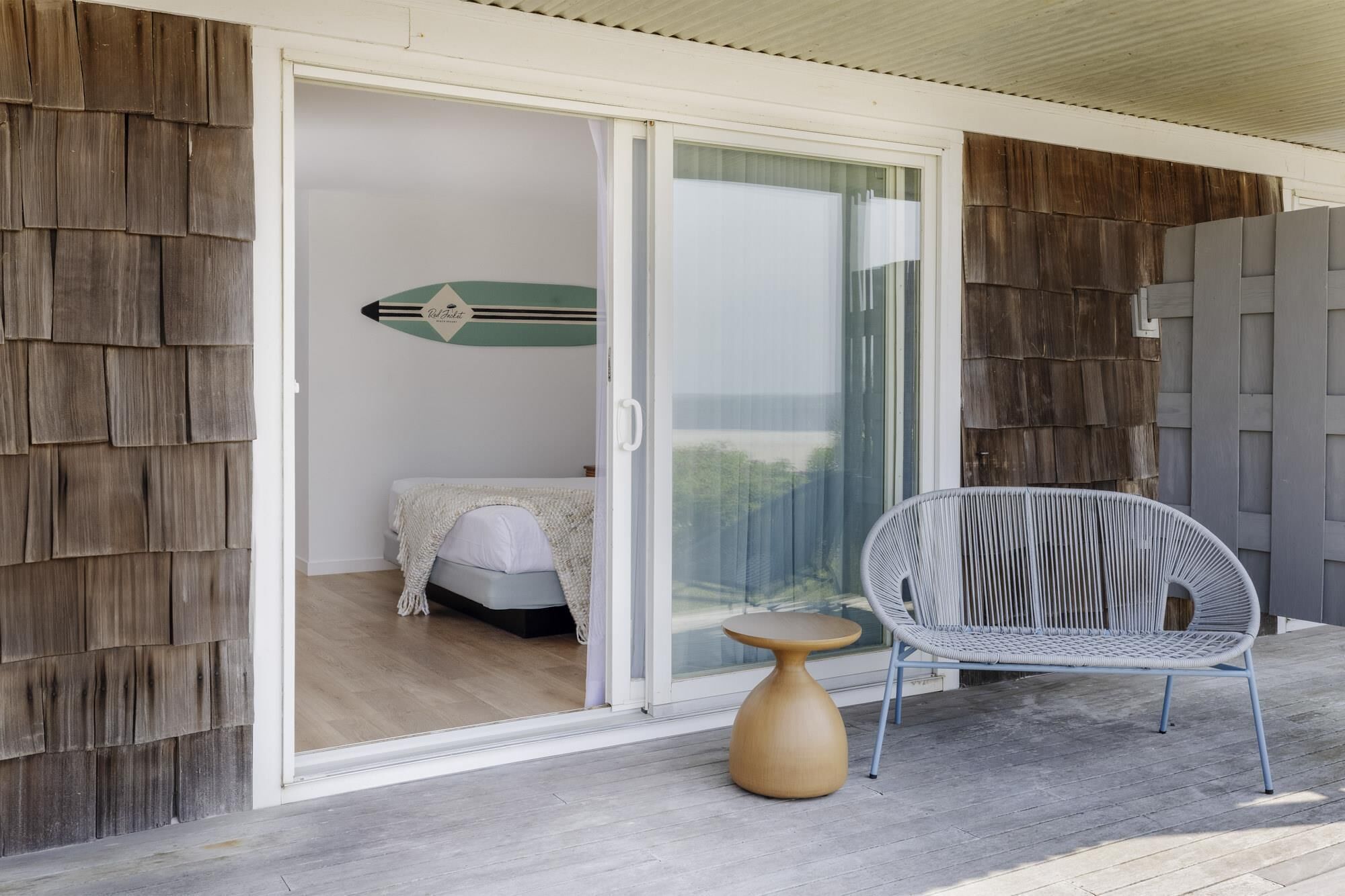 Ein Zimmer mit einer Schiebeglastür, die auf einen Balkon führt, einem Bett mit weißer Bettdecke, einem Korbstuhl und einem kleinen Holztisch.