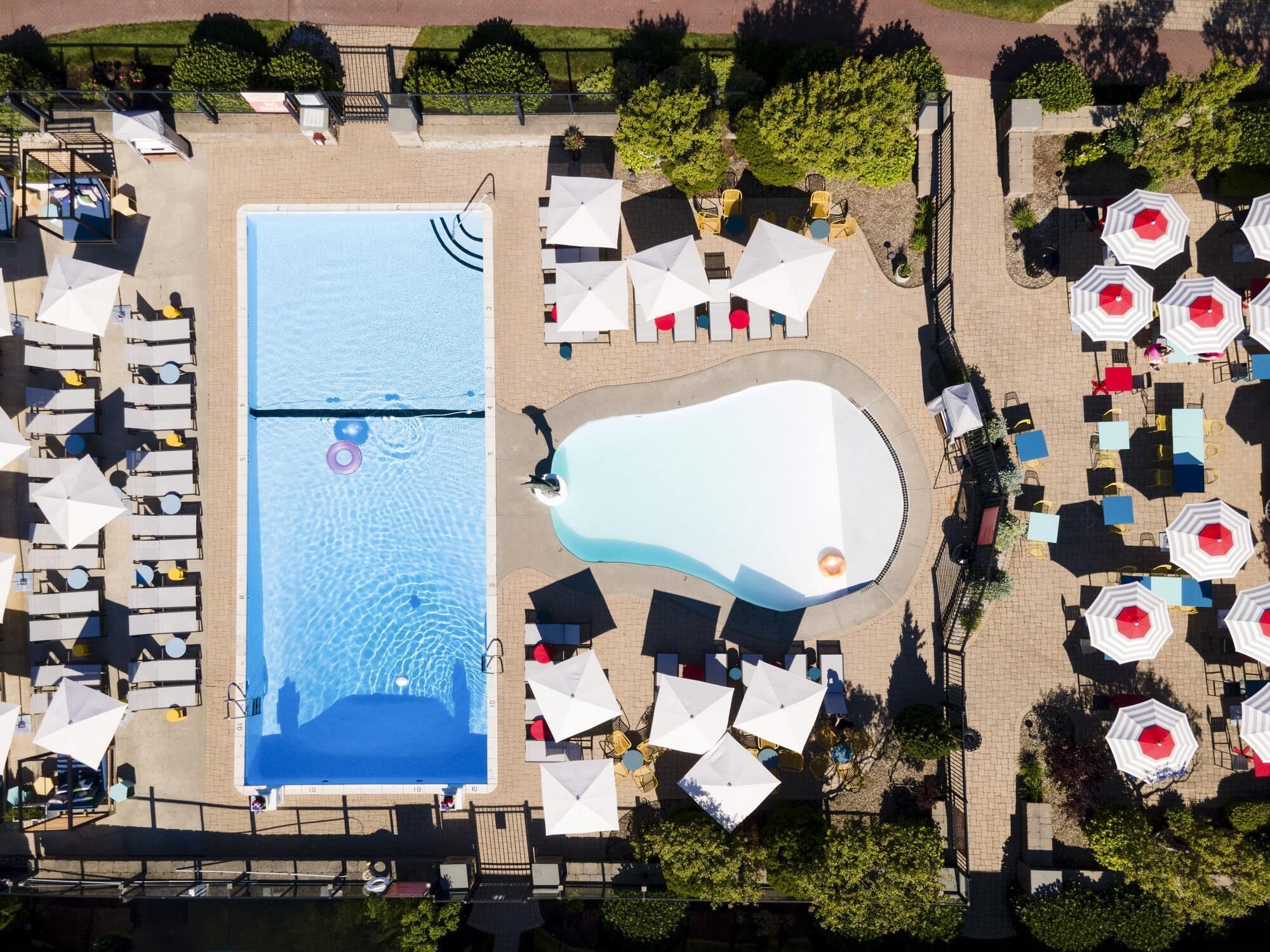 An aerial view of a hotel with a large rectangular pool, smaller circular pool, and multiple outdoor seating areas with umbrellas.