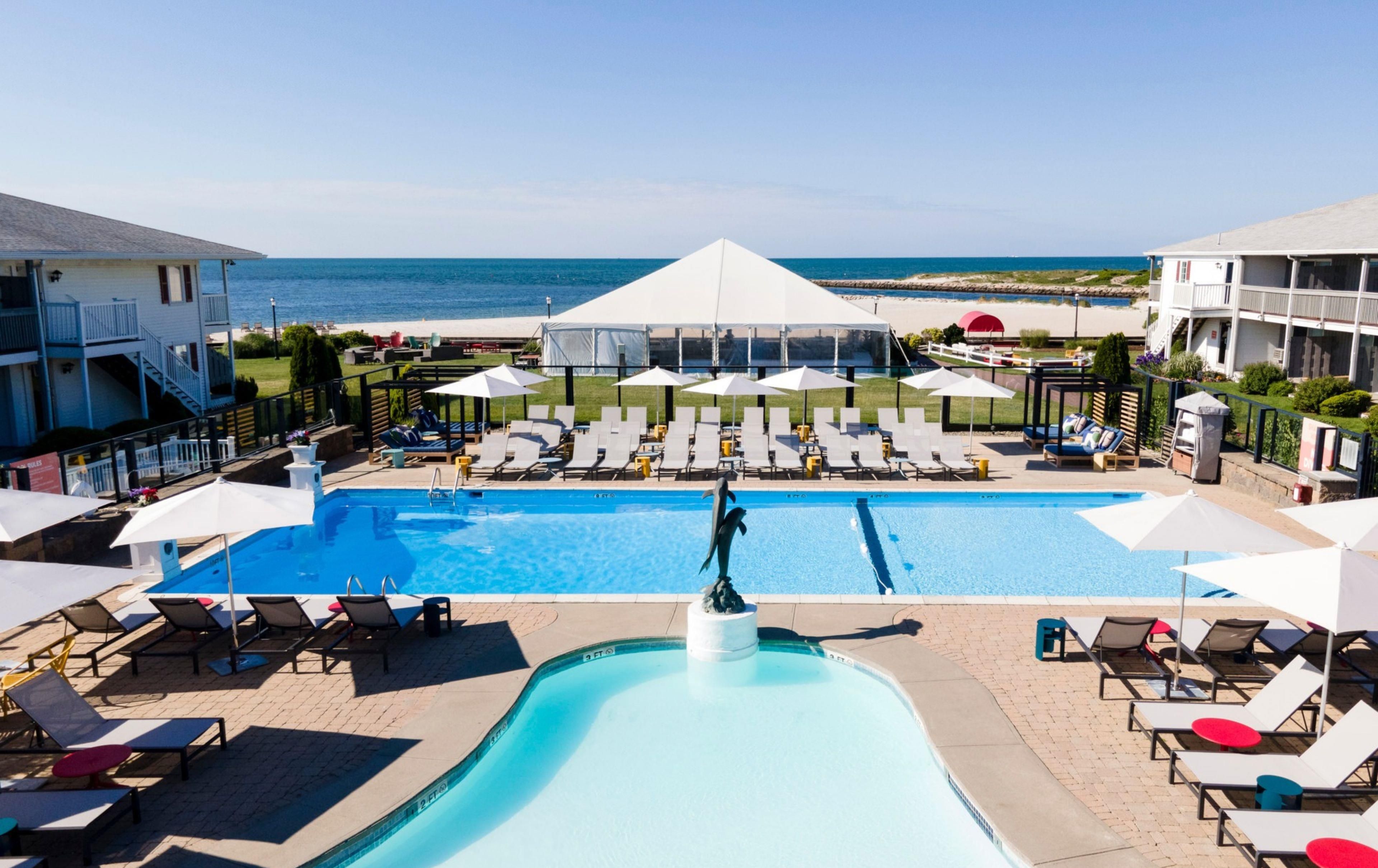 Una piscina con sillas de descanso y sombrillas, una zona cubierta y vista al mar.