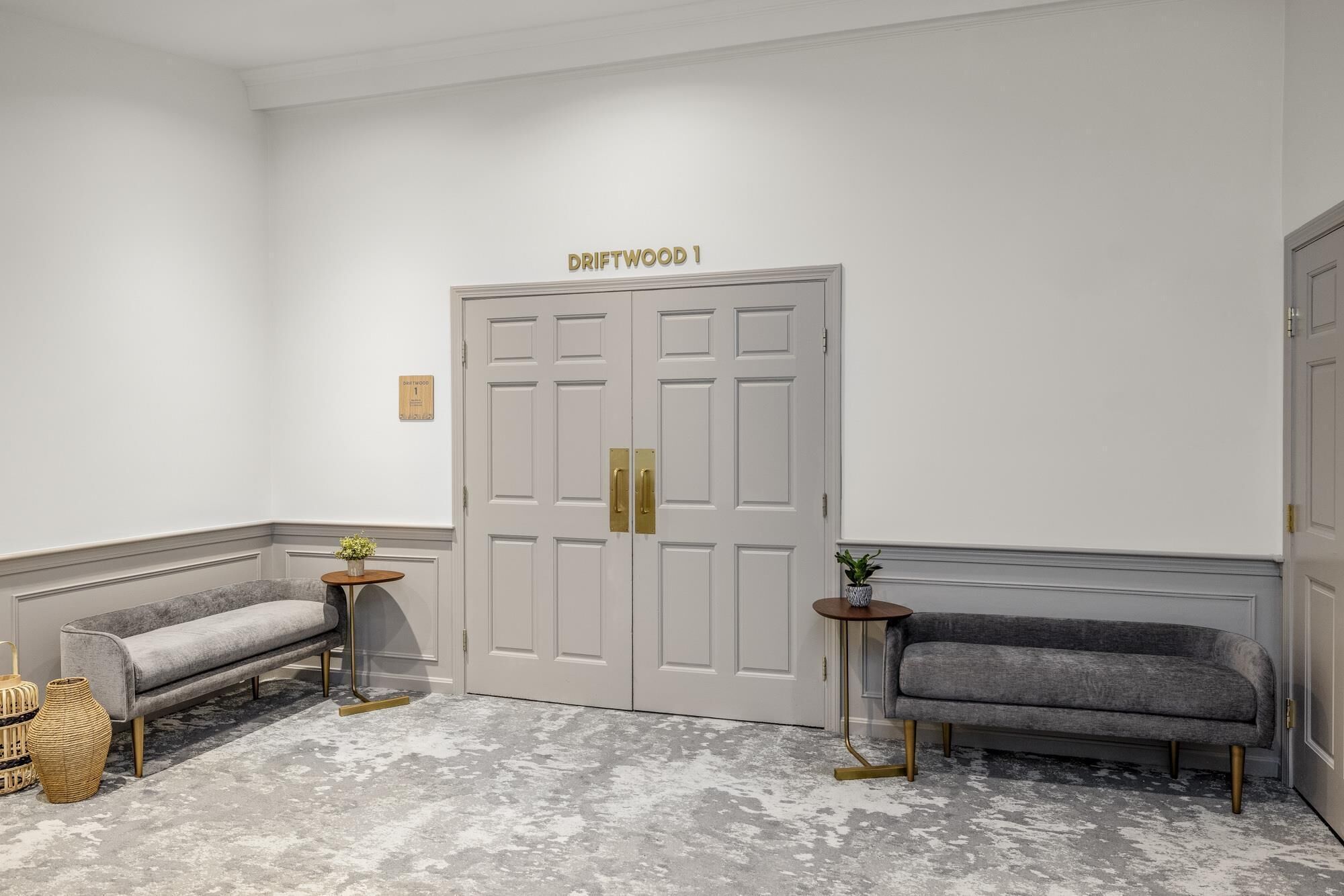 Um corredor com dois sofás cinza, uma porta branca com a inscrição "DRIFTWOOD 1" e um chão cinza texturizado.