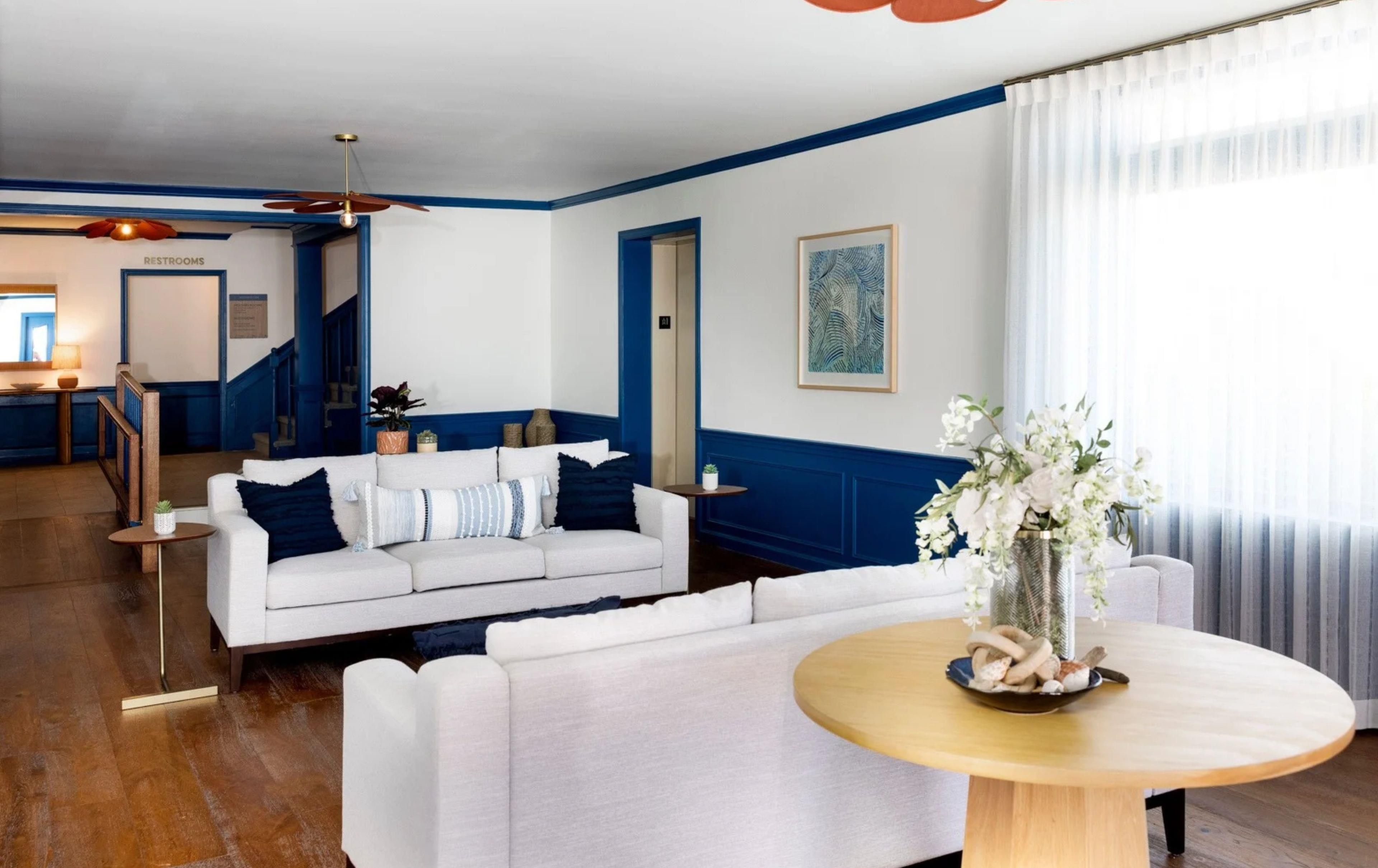 Une chambre d’hôtel avec un canapé blanc, une table ronde en bois, un tableau accroché au mur et un escalier menant à l’étage supérieur.