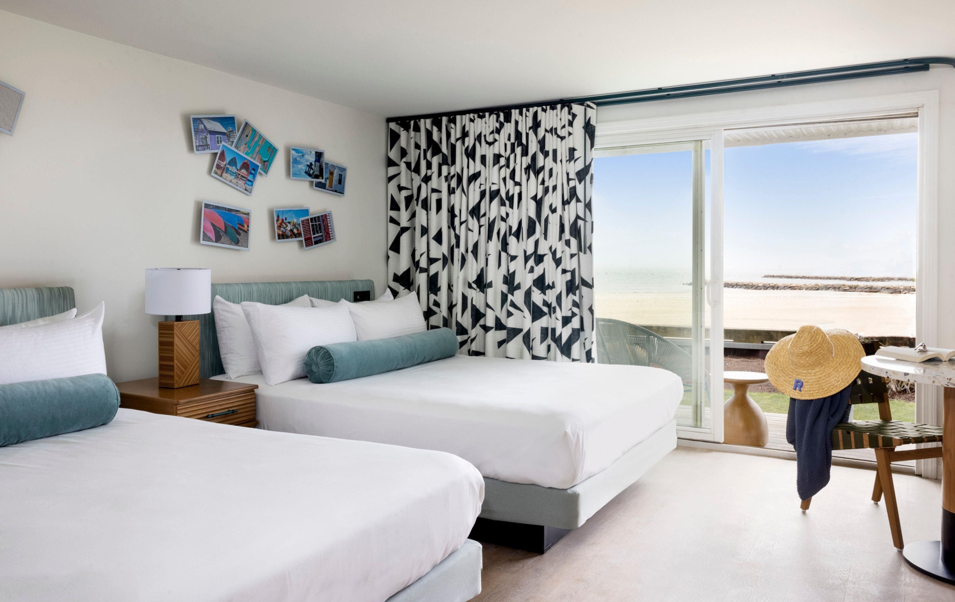 A hotel room with two beds, a large window with a view of the beach, and a straw hat on a chair.