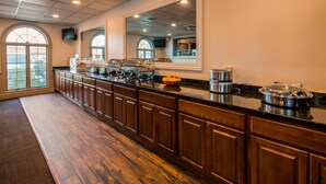 A long, dark wood-paneled counter with a variety of food items and appliances, large windows with arched tops, and a high ceiling with recessed lighting.