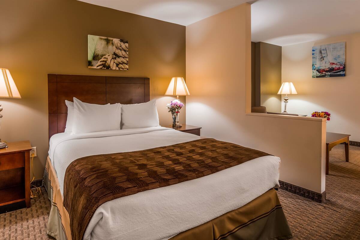 A hotel room with a large bed, two bedside tables with lamps, a wooden headboard, a painting on the wall, a desk with a chair, and a mirror.