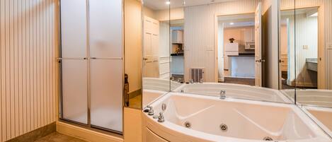 A modern bathroom with a large bathtub, a walk-in shower, and a mirror reflecting the kitchen area.