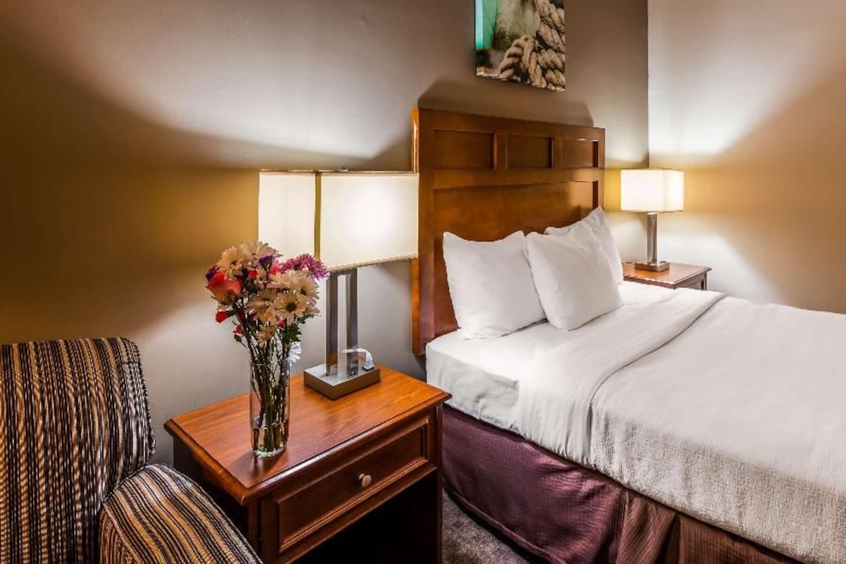 A hotel room with a bed, a nightstand with a lamp, a vase with flowers, and a chair.