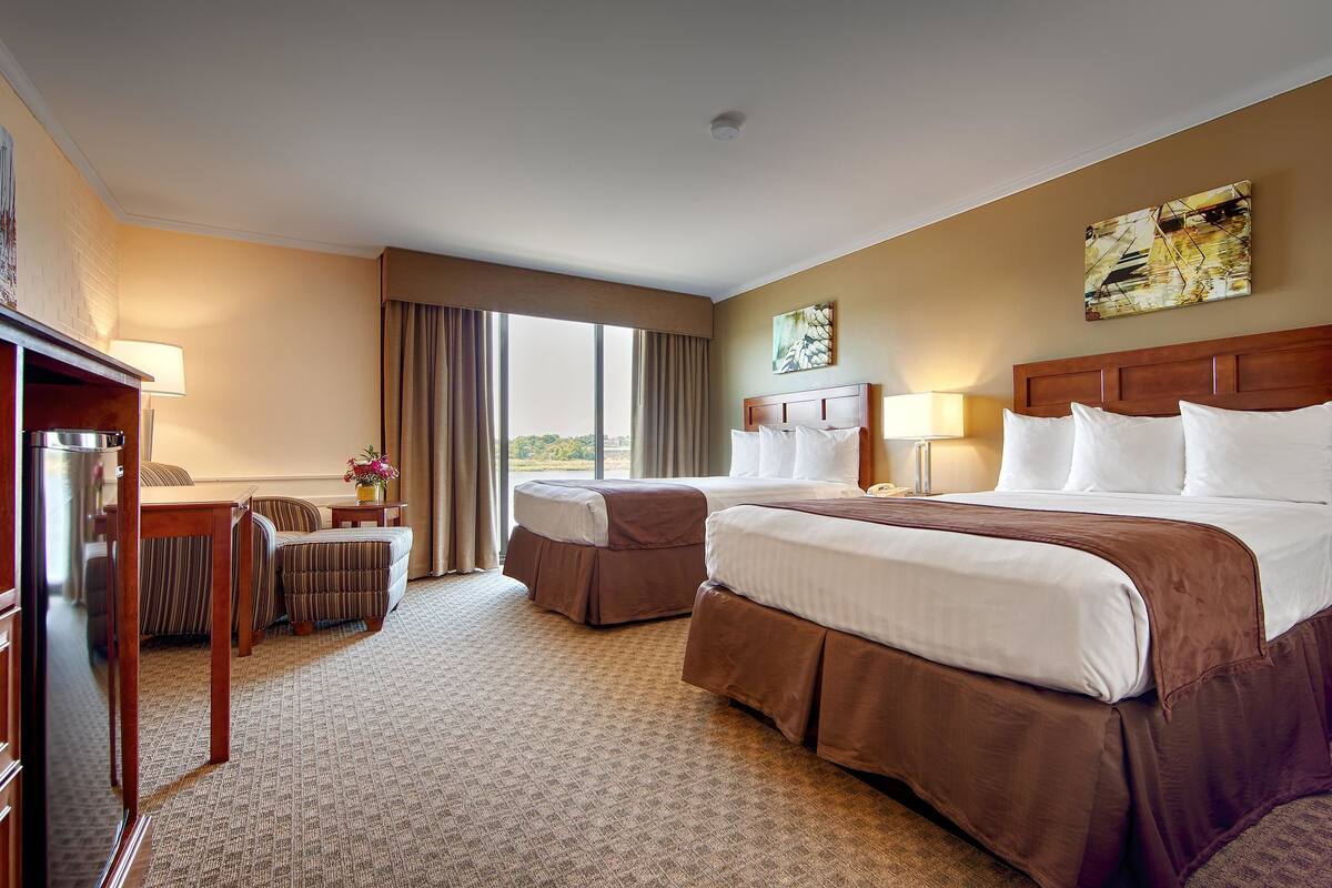 A hotel room with two beds, a desk, a chair, and a view of the outdoors.