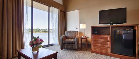 A hotel room with a large window, a television, a chair, a dresser, and a small table with a vase of flowers.