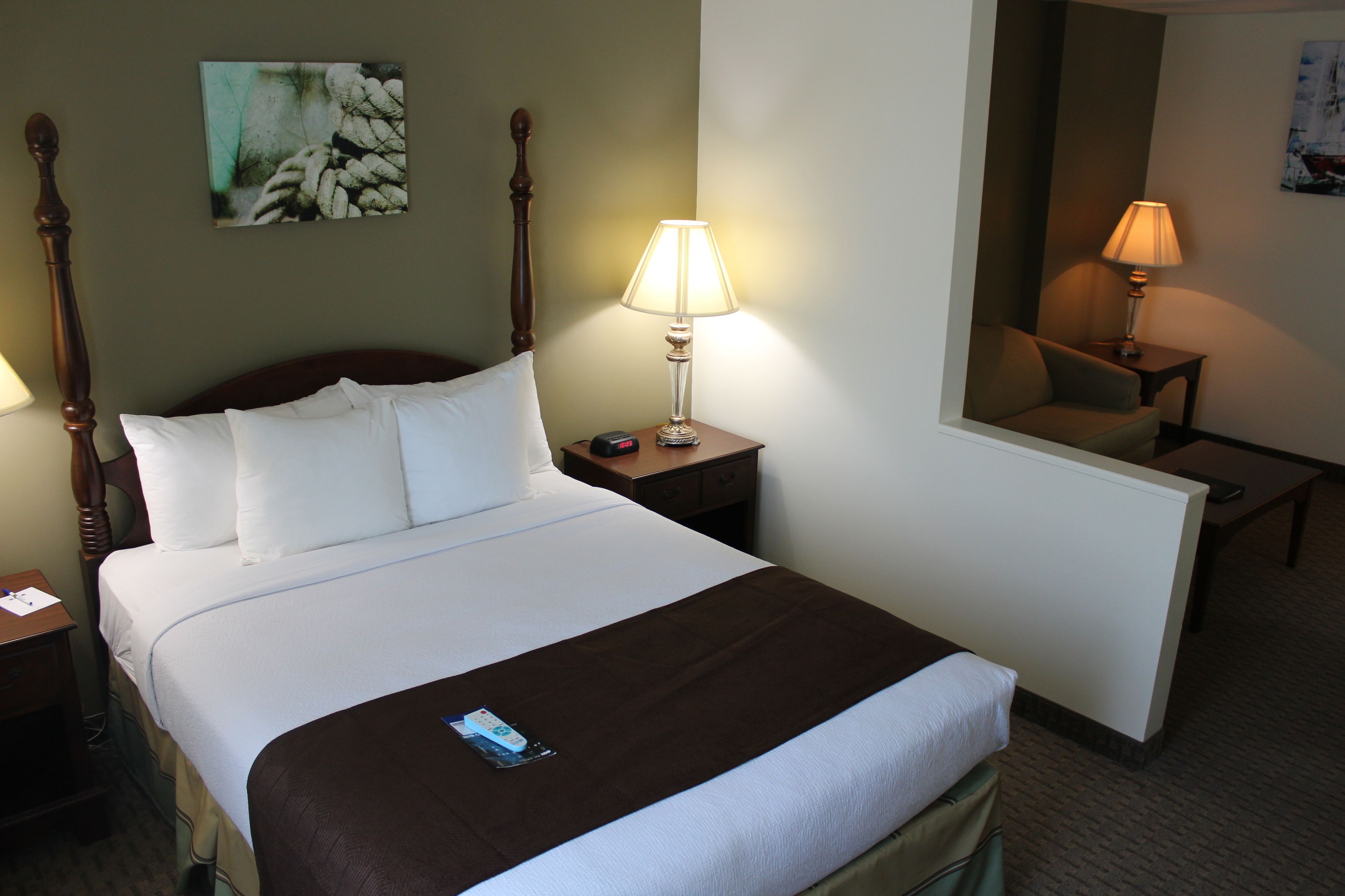 A neatly made bed with white linens and a brown throw, a bedside lamp, a nightstand, and a painting on the wall.