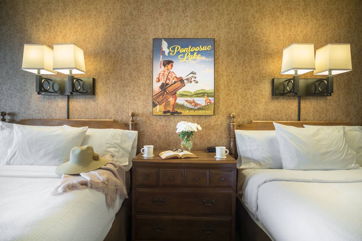 A hotel room with two beds, a nightstand with a lamp, a poster of Pontoosuc Lake, and a vase with flowers.