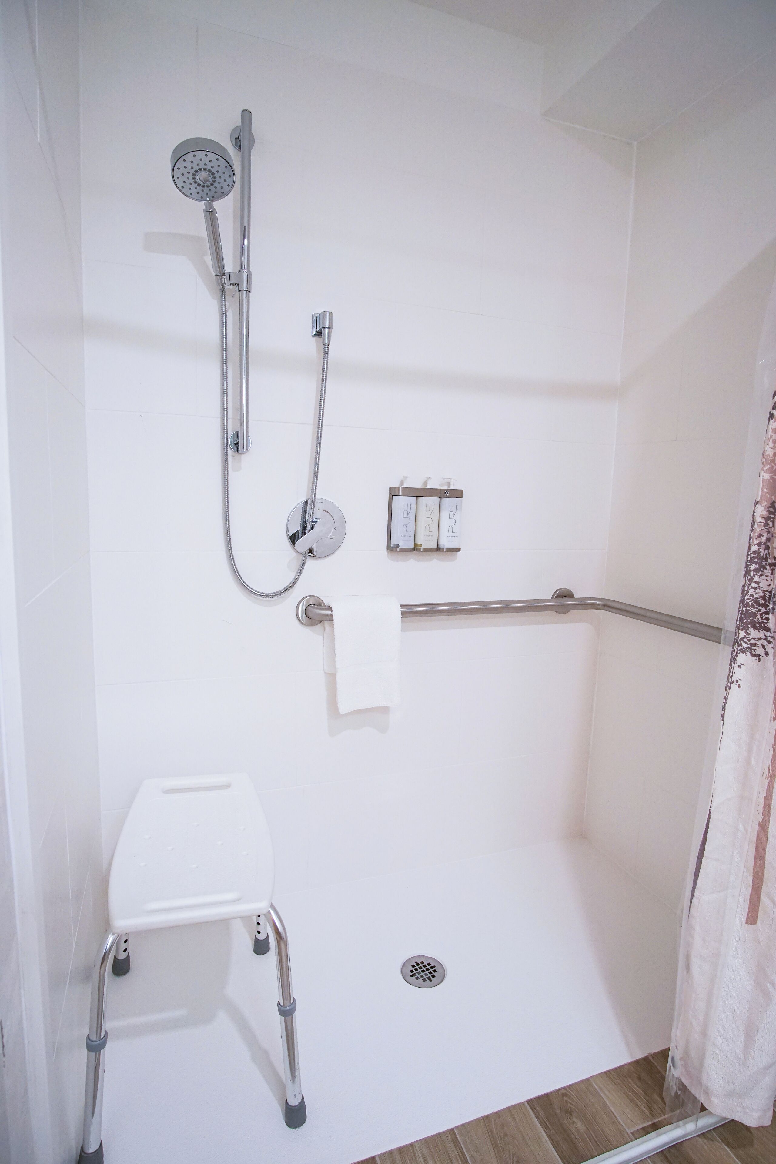 Zona de ducha con cabezal de ducha de mano, ducha de mano adicional, barra de apoyo, inodoro blanco y un taburete pequeño.