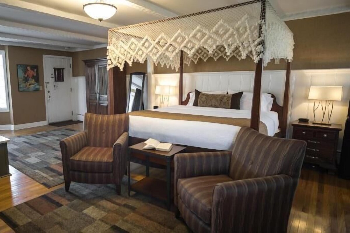 Ett hotellrum med en säng med himmelssäng, en sittgrupp med två fåtöljer, ett stort fönster och ett badrum med handfat.