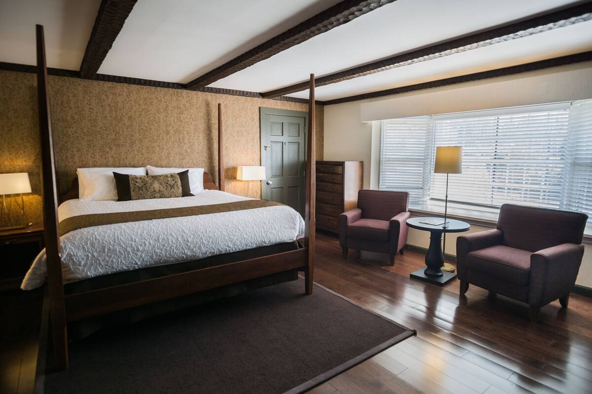 A four-poster bed with a wooden frame, a small round table, two armchairs, and a window with blinds.