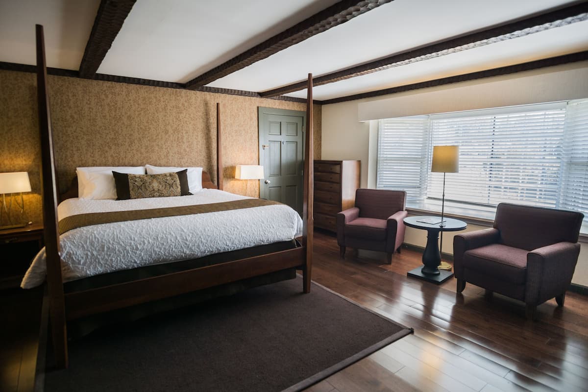 A four-poster bed with a wooden frame, a small round table, two armchairs, and a window with blinds.
