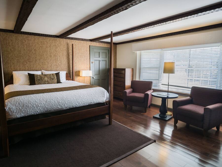 Una cama con dosel de madera, una mesita redonda pequeña, dos sillones y una ventana con persianas.