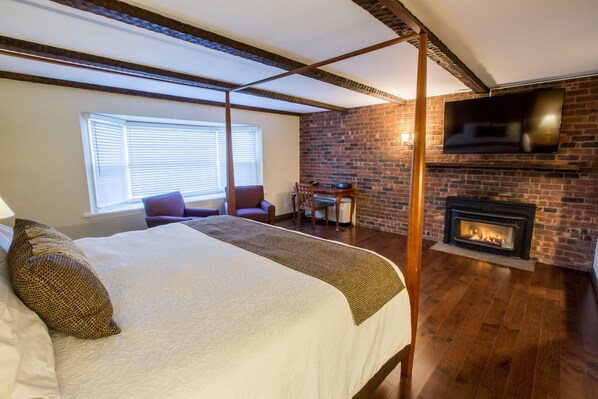 A bedroom with a large bed, a brick fireplace, a TV mounted on the wall, and a window with blinds. - The Yankee Inn (Lenox)
