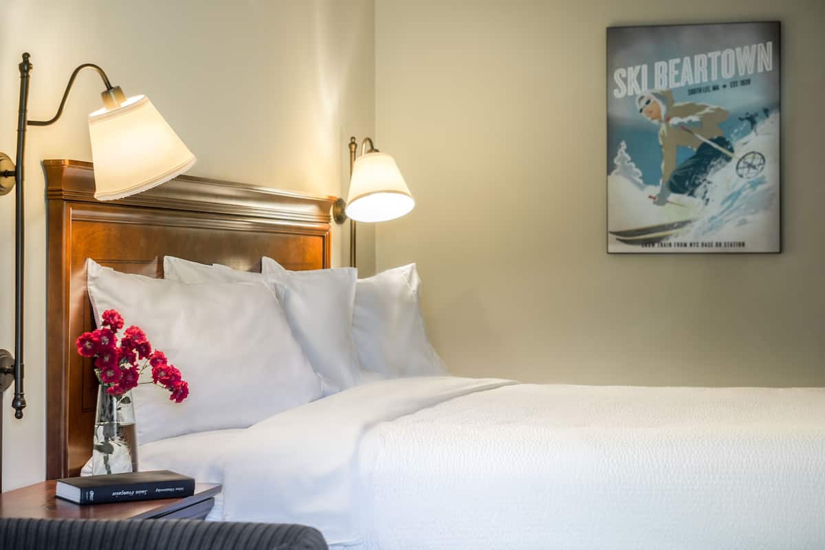 A hotel room with a wooden headboard, white bedding, a vase with red flowers, and a poster on the wall.