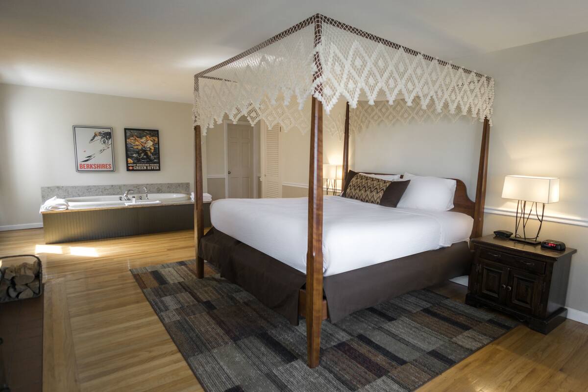 A four-poster canopy bed with a wooden frame and white bedding, a small bedside table with a lamp, and a large window with a view of the city.
