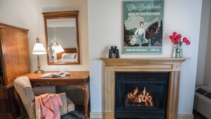 A cozy room with a wooden desk, a chair with a blanket, a fireplace with a fire, a mirror, and a poster on the wall. - The Yankee Inn (Lenox)