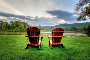 Two Adirondack chairs facing a river with a forested hill in the background.