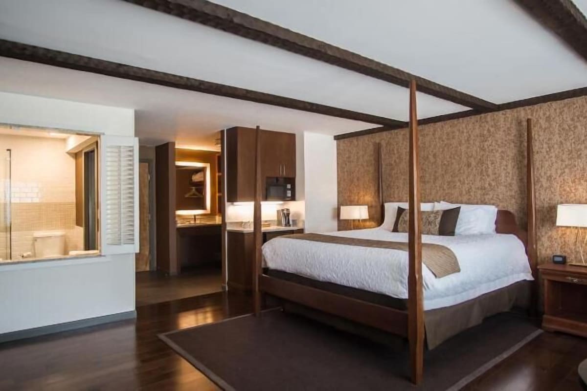 A four-poster bed with a wooden frame, a bathroom with a glass shower enclosure, and a small bedside table with a lamp.