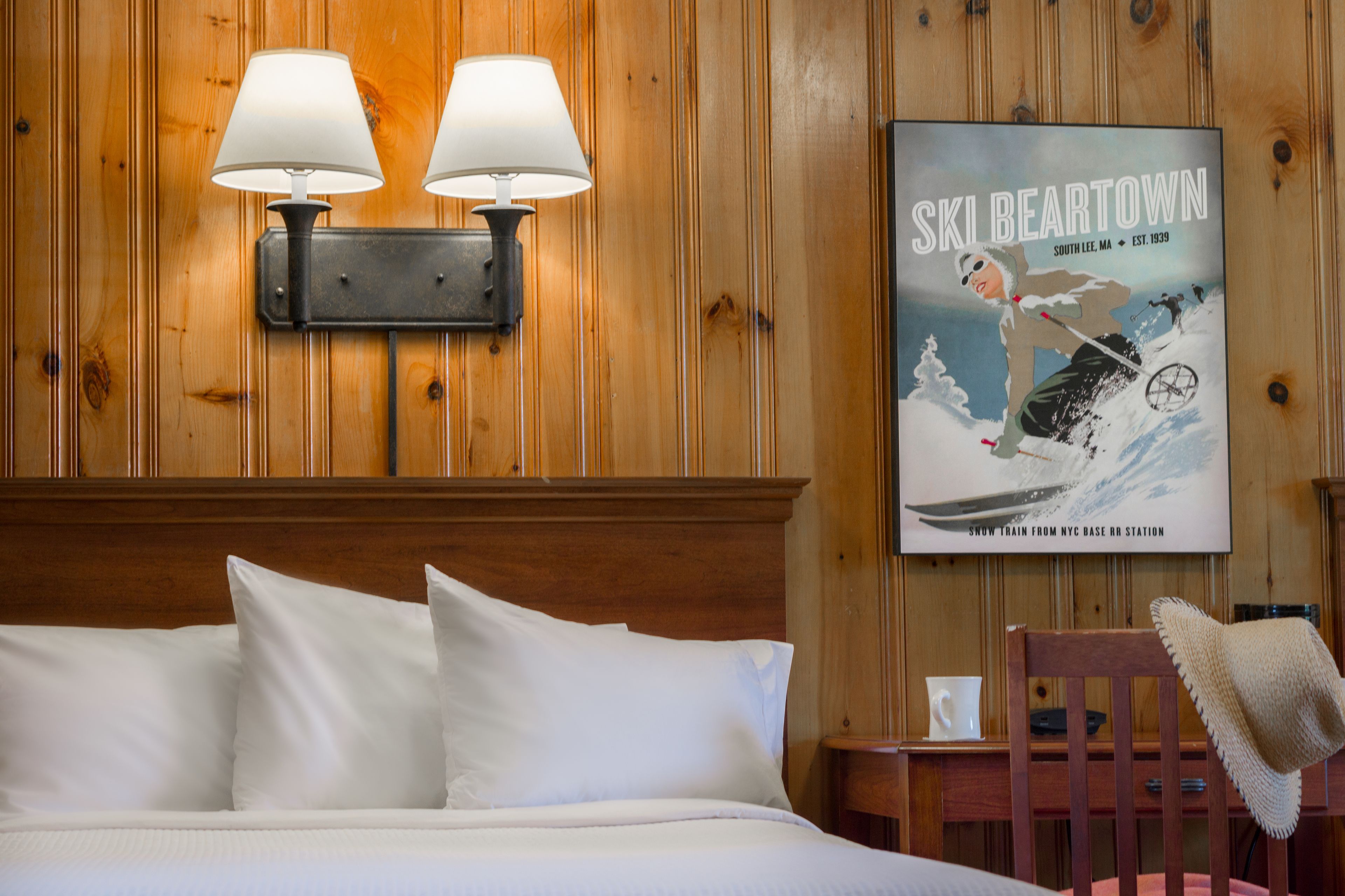 Quarto de hotel com parede revestida de madeira, cama com lençóis brancos, duas luminárias fixadas na parede, um cartaz com uma cena de esqui e uma pequena mesa com um chapéu em cima.