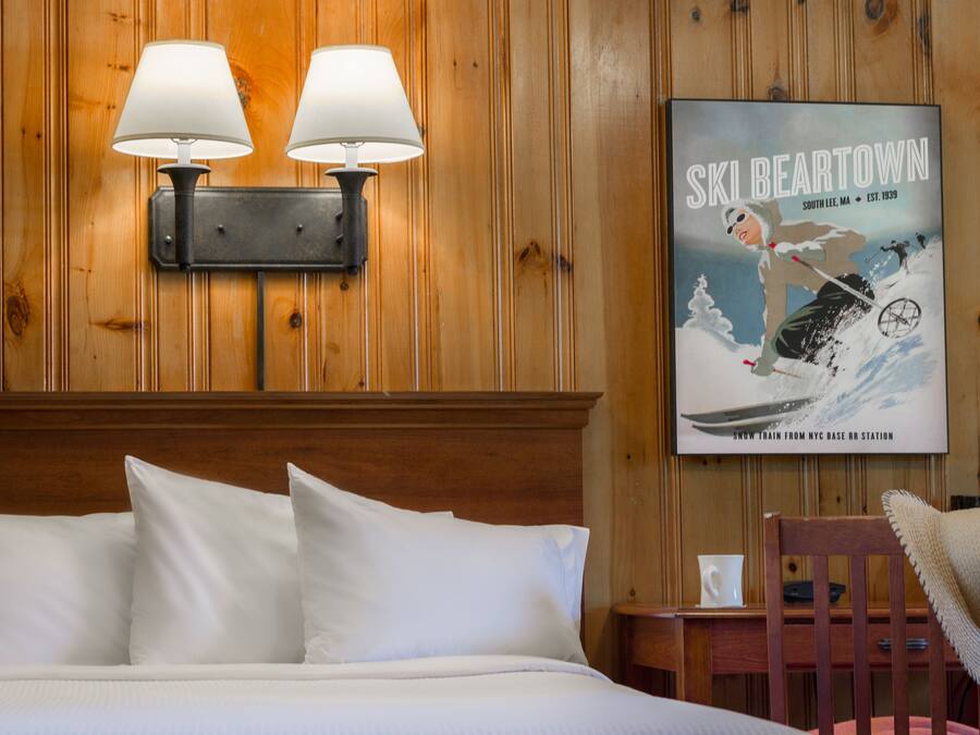 Habitación de hotel con pared de paneles de madera, cama con ropa blanca, dos lámparas fijadas a la pared, un cartel con una escena de esquí y una mesita con un sombrero encima.