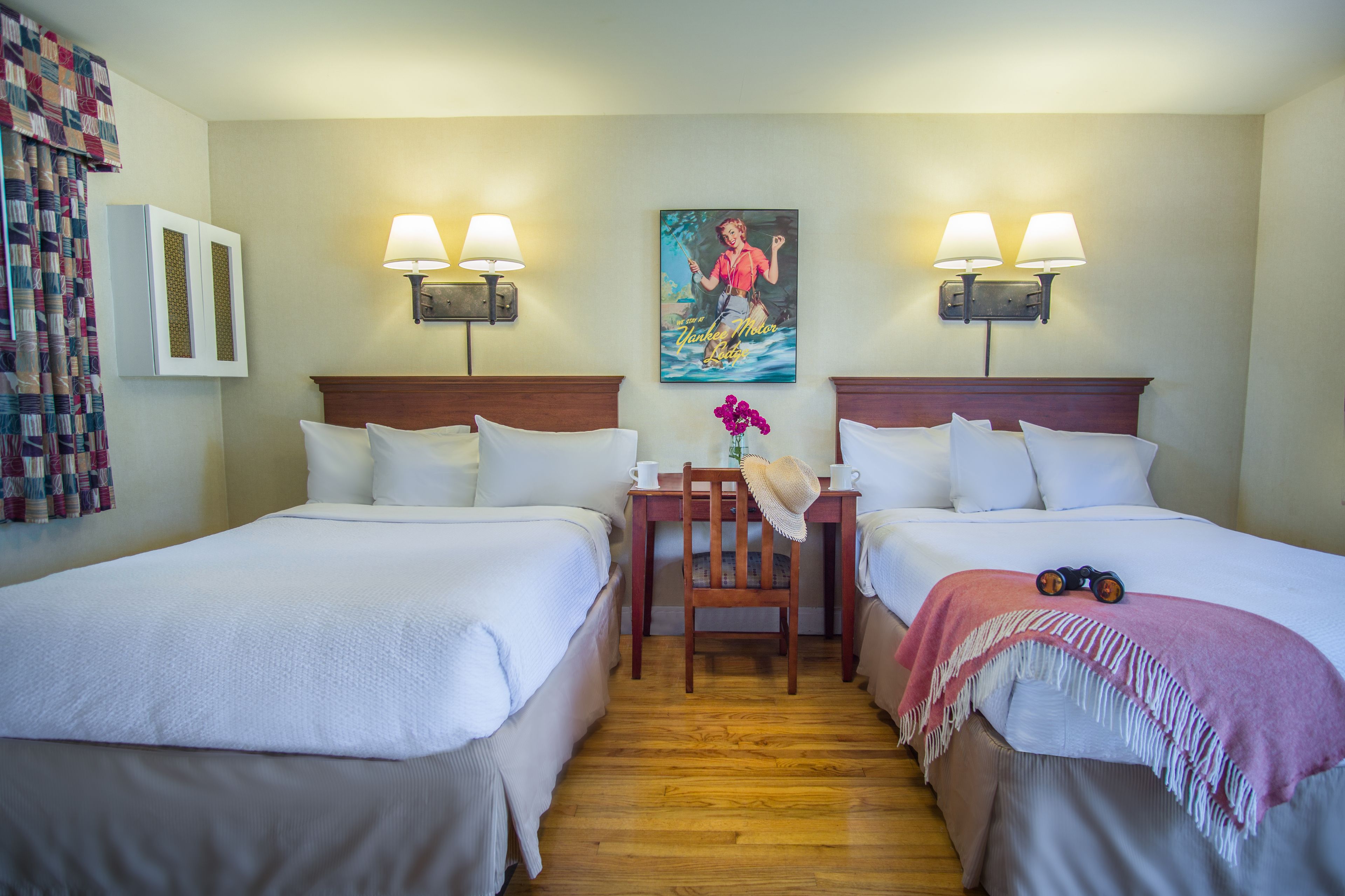 A hotel room with two beds, a wooden table, a chair, and a painting on the wall.