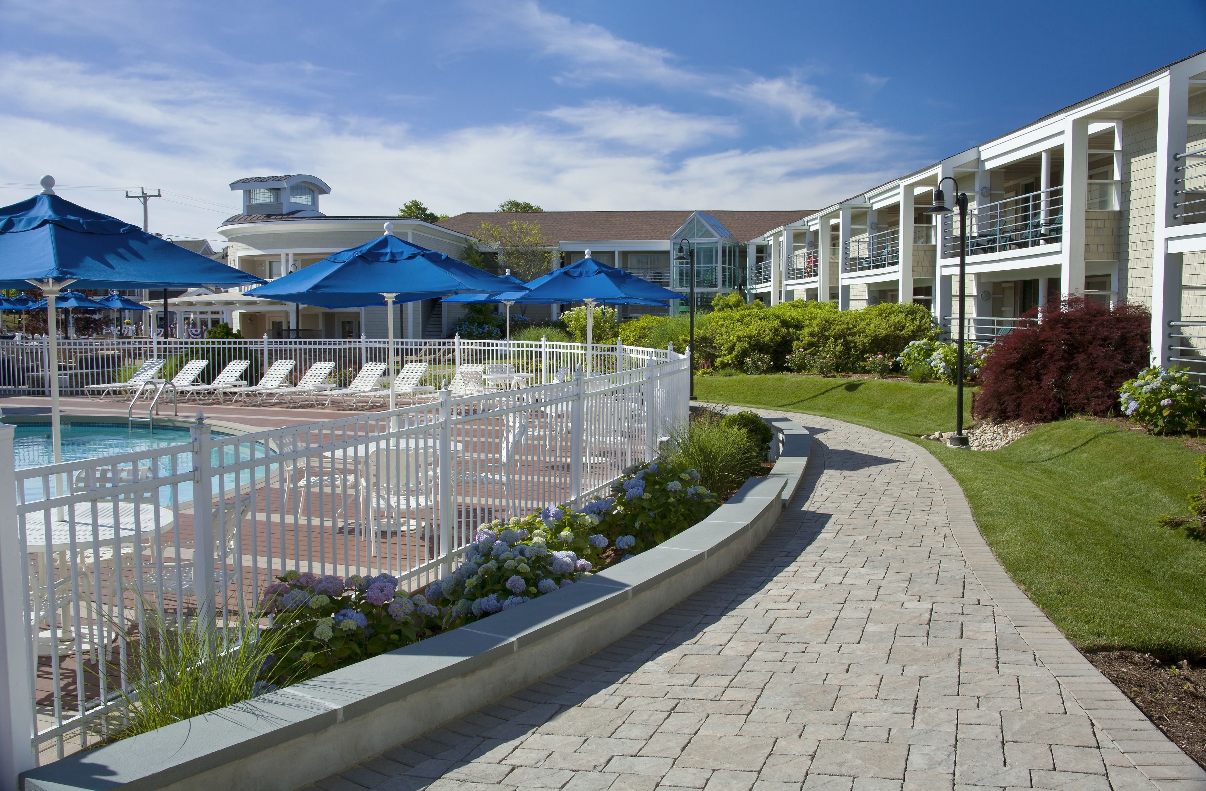 A pool area with lounge chairs and umbrellas, a brick pathway, and a building with balconies.